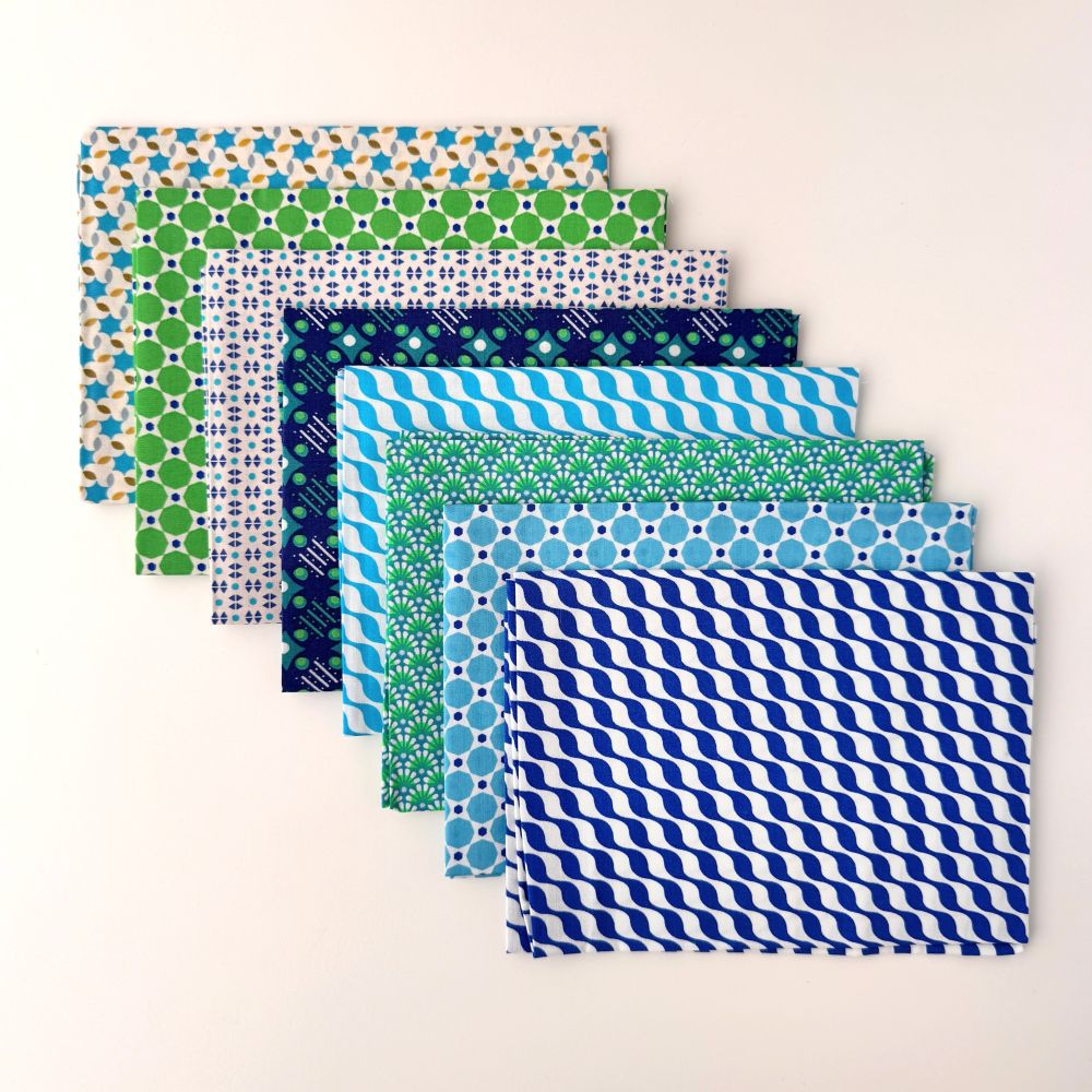 A packaged fabric bundle labeled Petit Pan Fabrics, showing a set of patterned fabric swatches wrapped with a white paper band. The fabric has a repeating geometric pattern in blue, tan, and white. Kifkif Overseas Petit Pan Fabric Bundle | 8-Piece Coupon Kit
