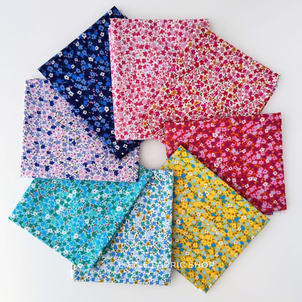 Six folded fabric squares with different floral and ditsy print patterns in various colors, arranged in a fan-like layout with a small white ball in the center. The patterns include blue, pink, purple, teal, and yellow on white backgrounds. Petite Prints | August Ellery | Half Yard Bundle