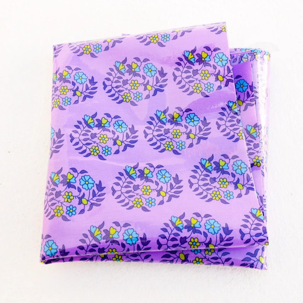 A folded purple cotton fabric with a floral pattern, wrapped in a light blue paper band labeled 'Petit Pan'. Oilcloth Coupon | Petit Pan | Shalin Lilac